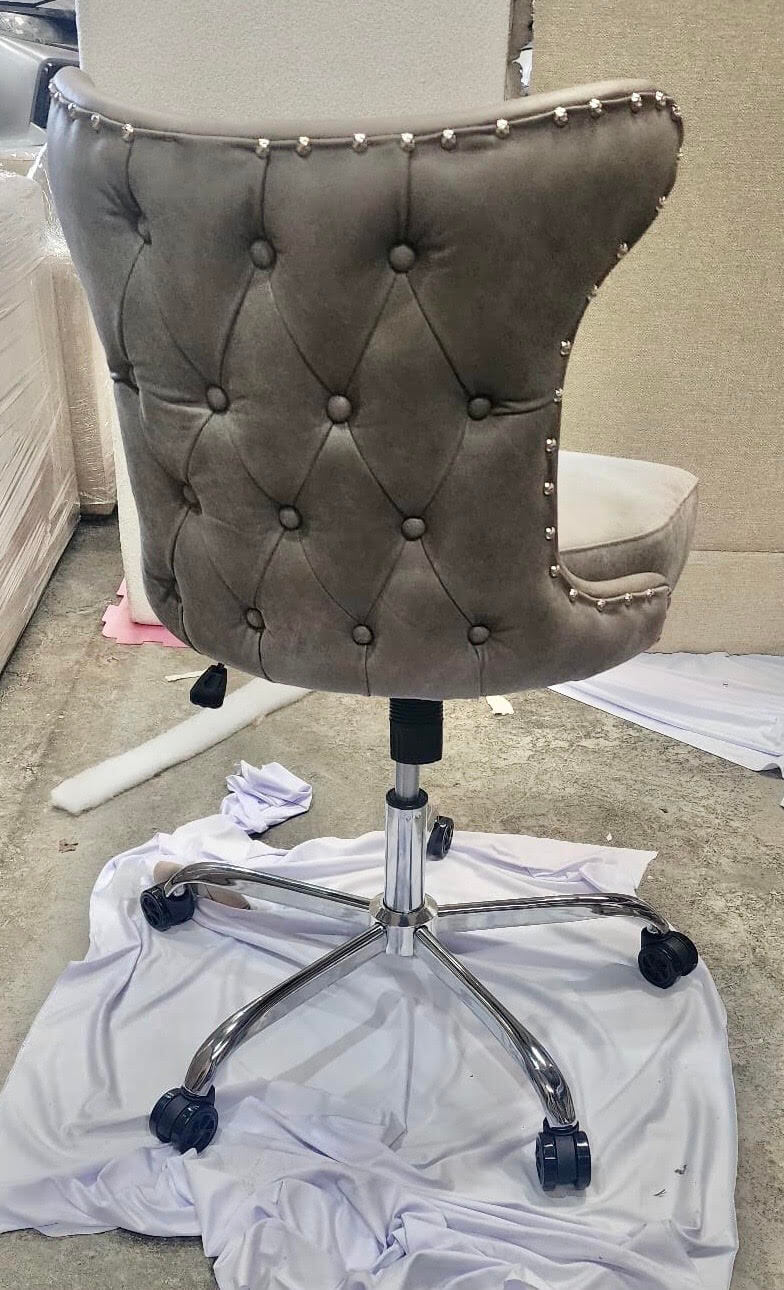 Back of a grey button-tufted leather poker chair with stud trim on a chrome swivel base with castors