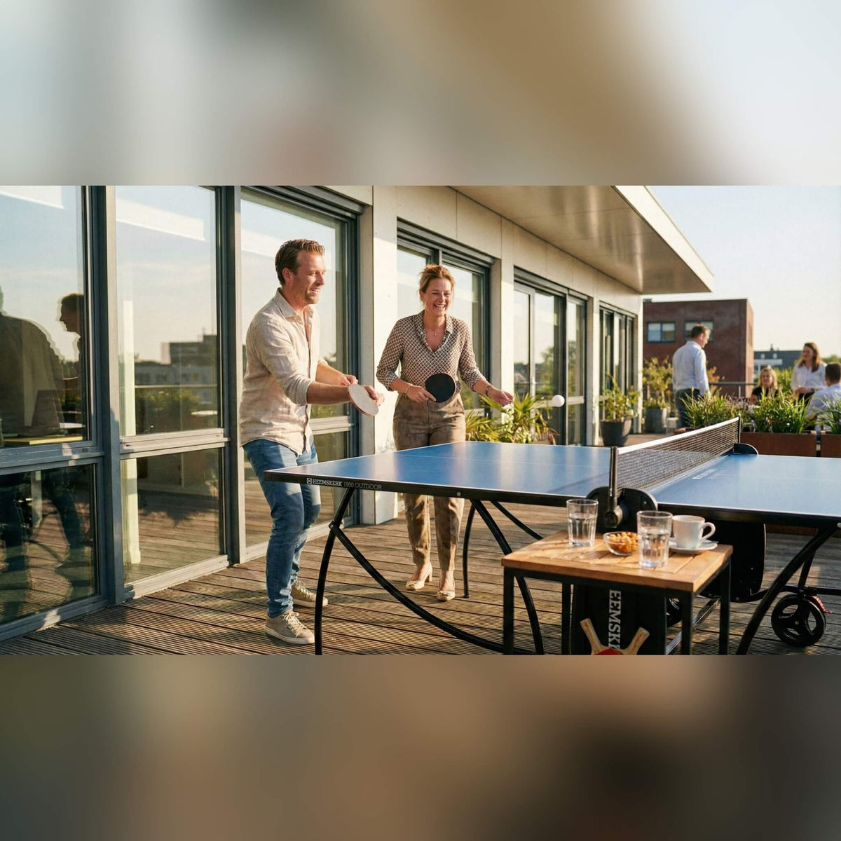Couple playing on a blue Heemskerk 1900 outdoor table tennis table on an office terrace
