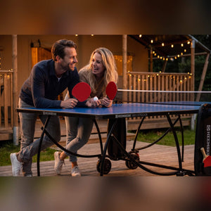 Couple with bats leaning on a blue Heemskerk 1900 outdoor table tennis table at dusk