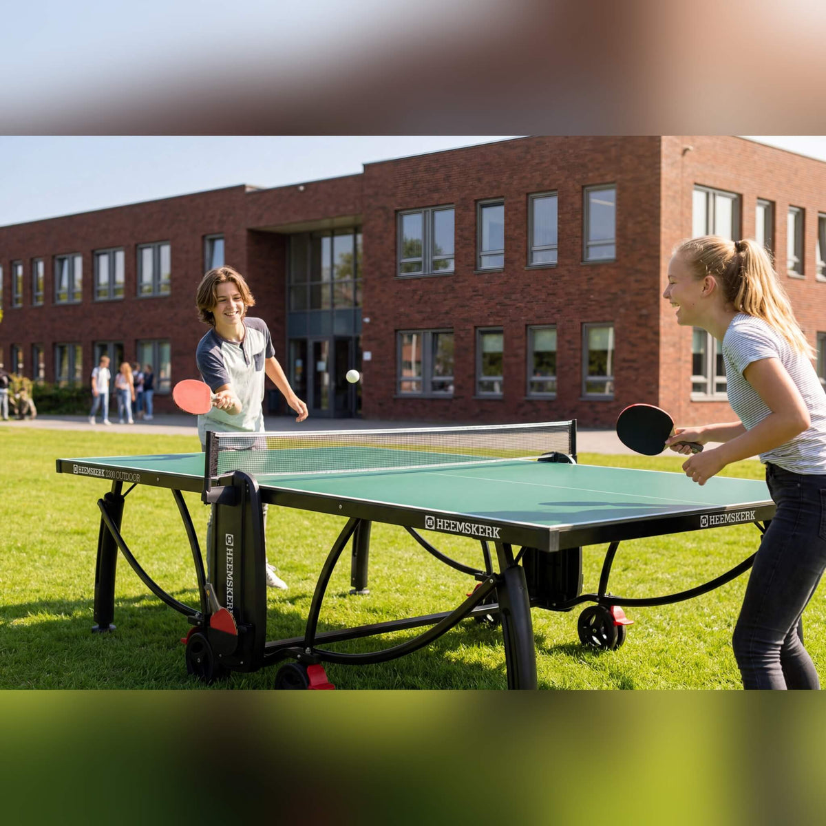 Two girls playing on a green Heemskerk 2300 outdoor table tennis table on a school lawn