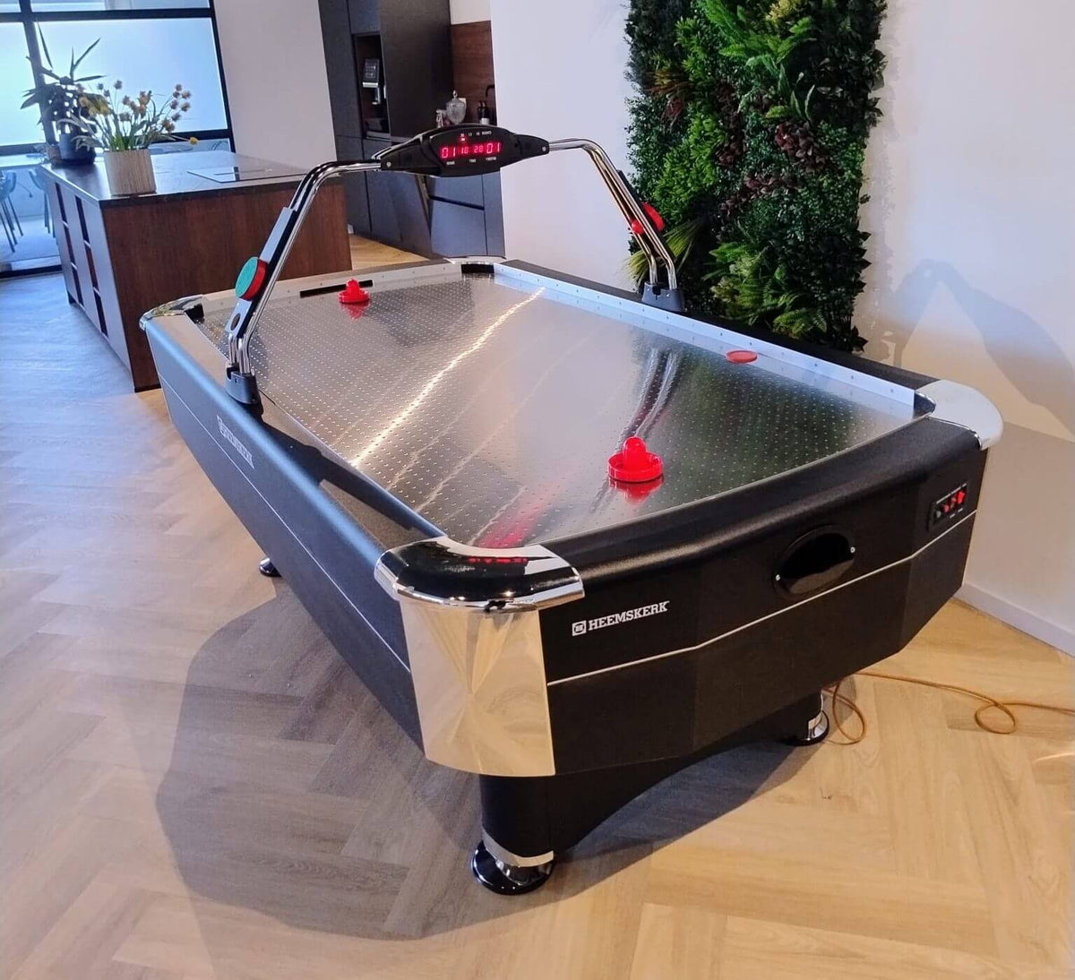 Heemskerk Navona air hockey table with a red pusher on the metal playfield in a lounge with a plant wall