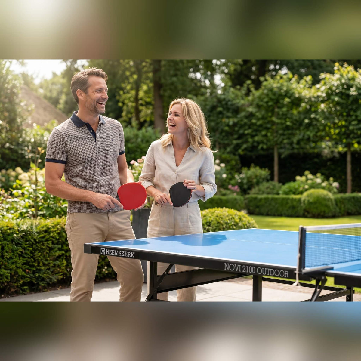 Couple holding bats beside a Heemskerk Novi 2100 outdoor table tennis table in a garden