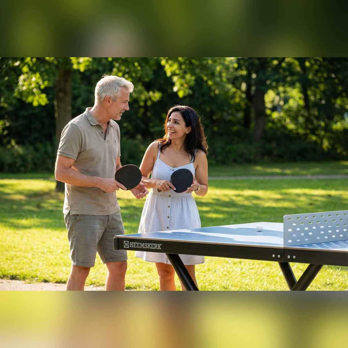 Couple holding bats beside a Heemskerk Tango outdoor table tennis table in a park