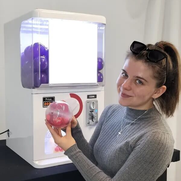 A woman holding a capsule beside a counter-top gashapon machine
